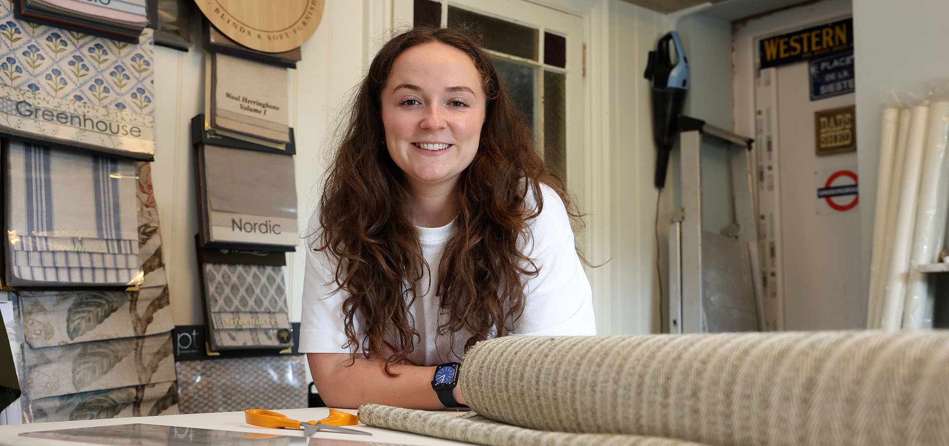 Lowri standing by a sewing table with fabric samples hanging on the wall behind